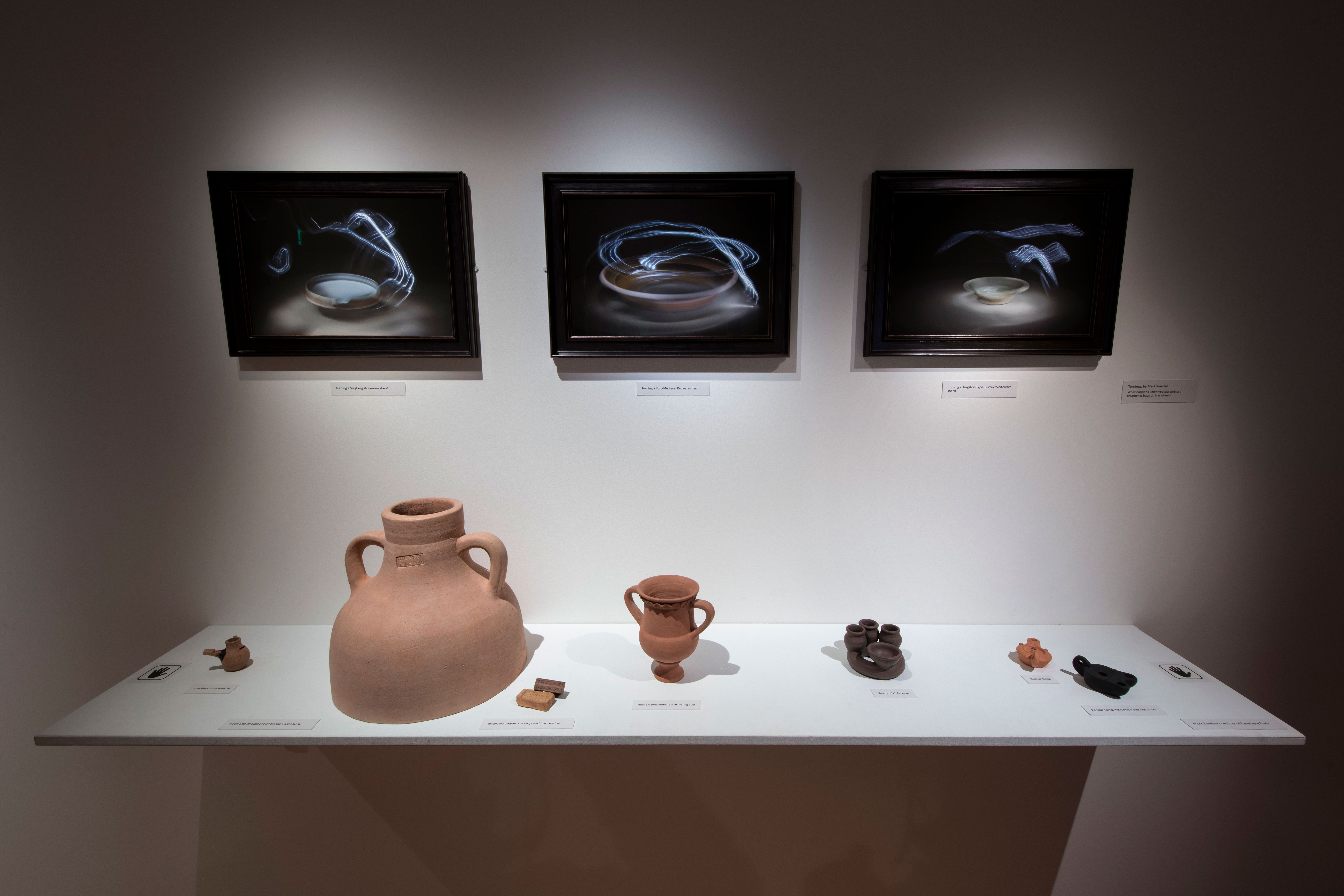 A photo of 3 long exposure photographs taken by Mark Sowden mounted on the gallery wall. Below the photographs are a selection pottery artefacts.